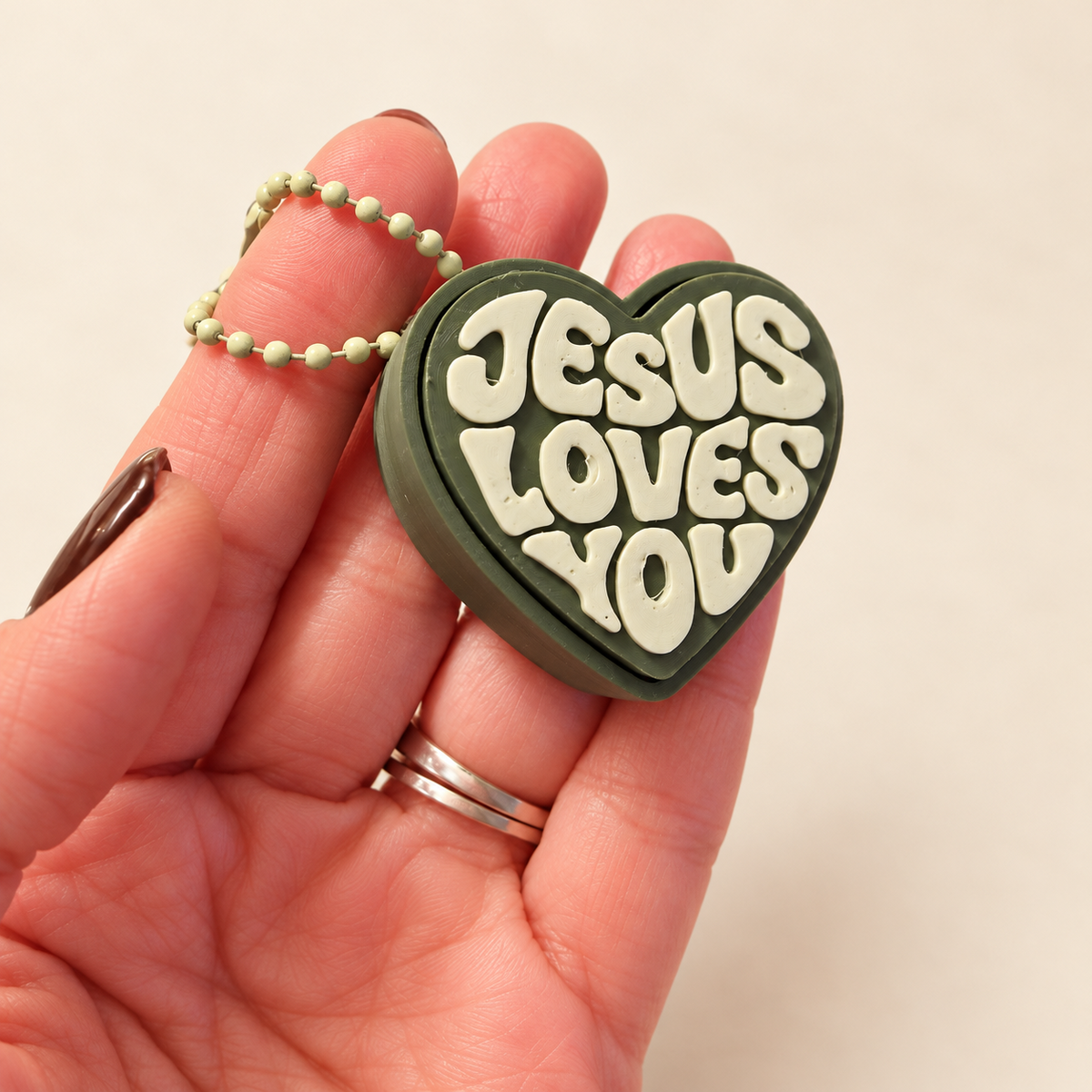 Olive green heart-shaped Jesus Loves You fidget clicker with cream lettering and a ball chain.